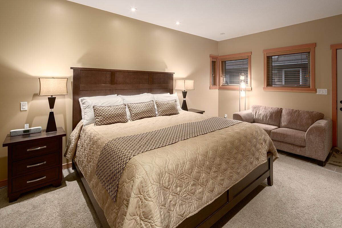 Master bedroom with king bed, taupe bedding, two night tables with lamps and couch to the right.