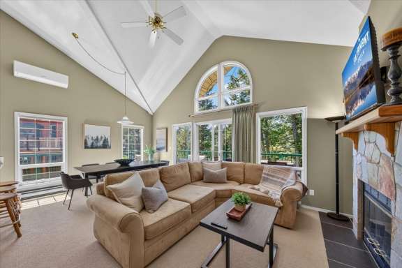 Open-concept living room with vaulted ceilings, a stone fireplace, large sectional sofa, dining area, and expansive windows overlooking the surrounding trees and deck in a Fairmont Creek vacation rental