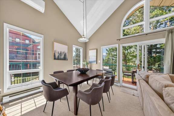 Bright dining area with seating for eight, vaulted ceilings, large window, and sliding doors leading to a private deck overlooking the golf course and surrounding trees in a Fairmont Creek vacation rental