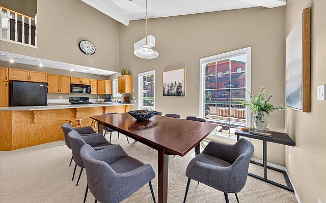 Open-concept living room with vaulted ceilings, a stone fireplace, large sectional sofa, dining area, and expansive windows overlooking the surrounding trees and deck in a Fairmont Creek vacation rental