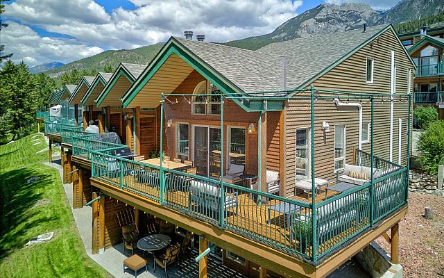 Open-concept living room with vaulted ceilings, a stone fireplace, large sectional sofa, dining area, and expansive windows overlooking the surrounding trees and deck in a Fairmont Creek vacation rental