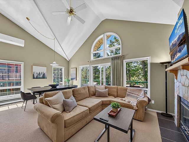 Open-concept living room with vaulted ceilings, a stone fireplace, large sectional sofa, dining area, and expansive windows overlooking the surrounding trees and deck in a Fairmont Creek vacation rental