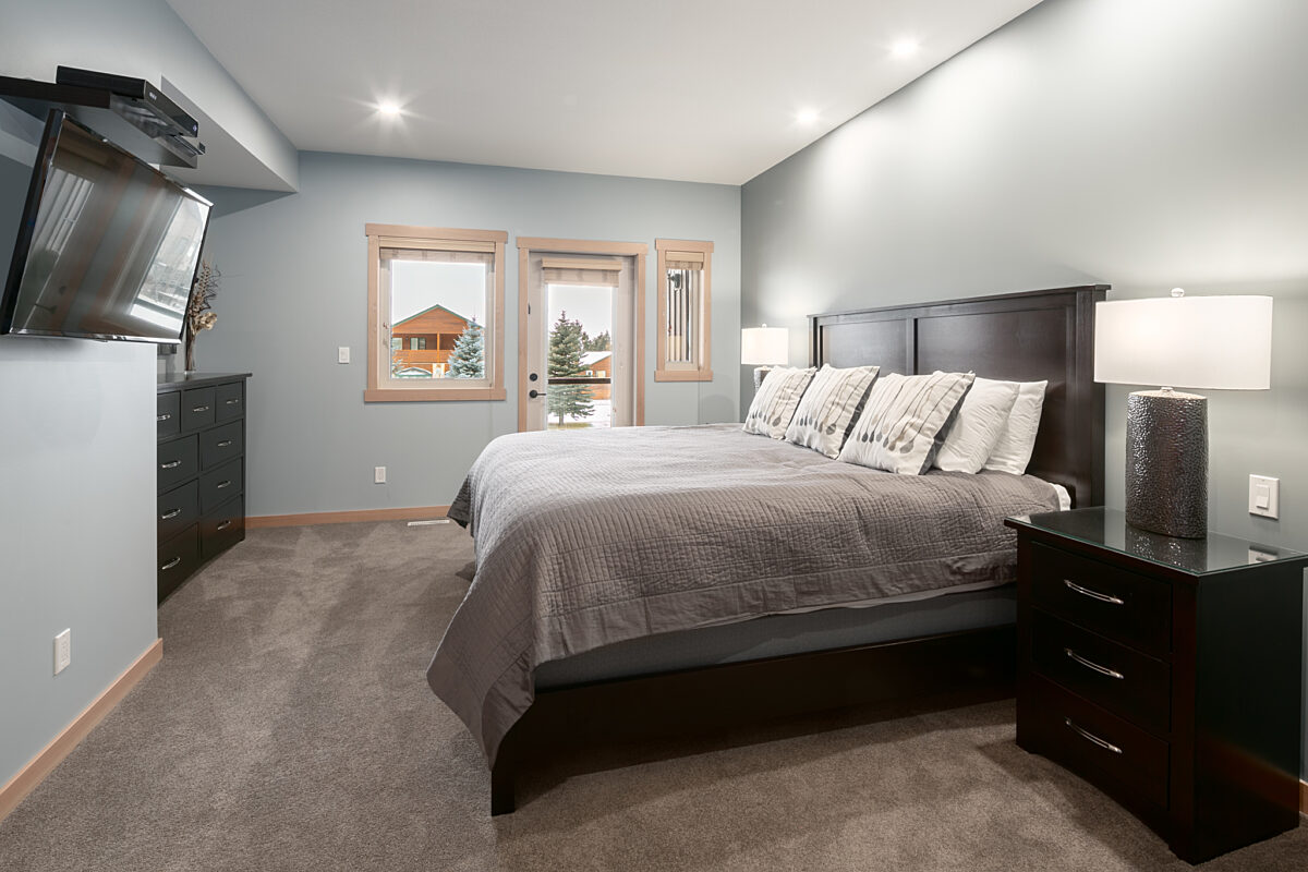 Queen bedroom with grey bedding, overhead lighting, two bedside tables, and windows to the left.