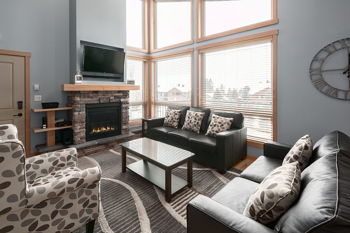 Large living room with windows to the right. Television above, two couches, one chair and coffee table.