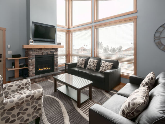 Large living room with windows to the right. Television above, two couches, one chair and coffee table.