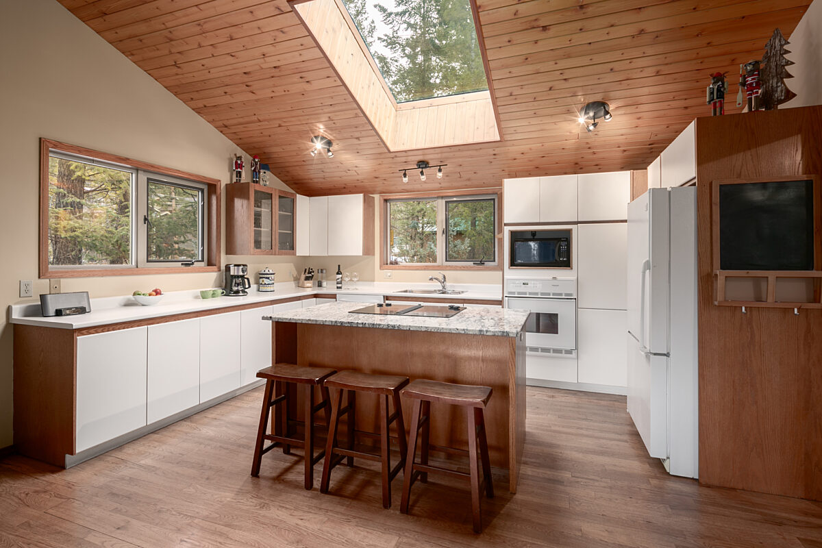 Large open concept kitchen with white cabinets and large windows. Kitchen island in the middle of the room with three barstools.