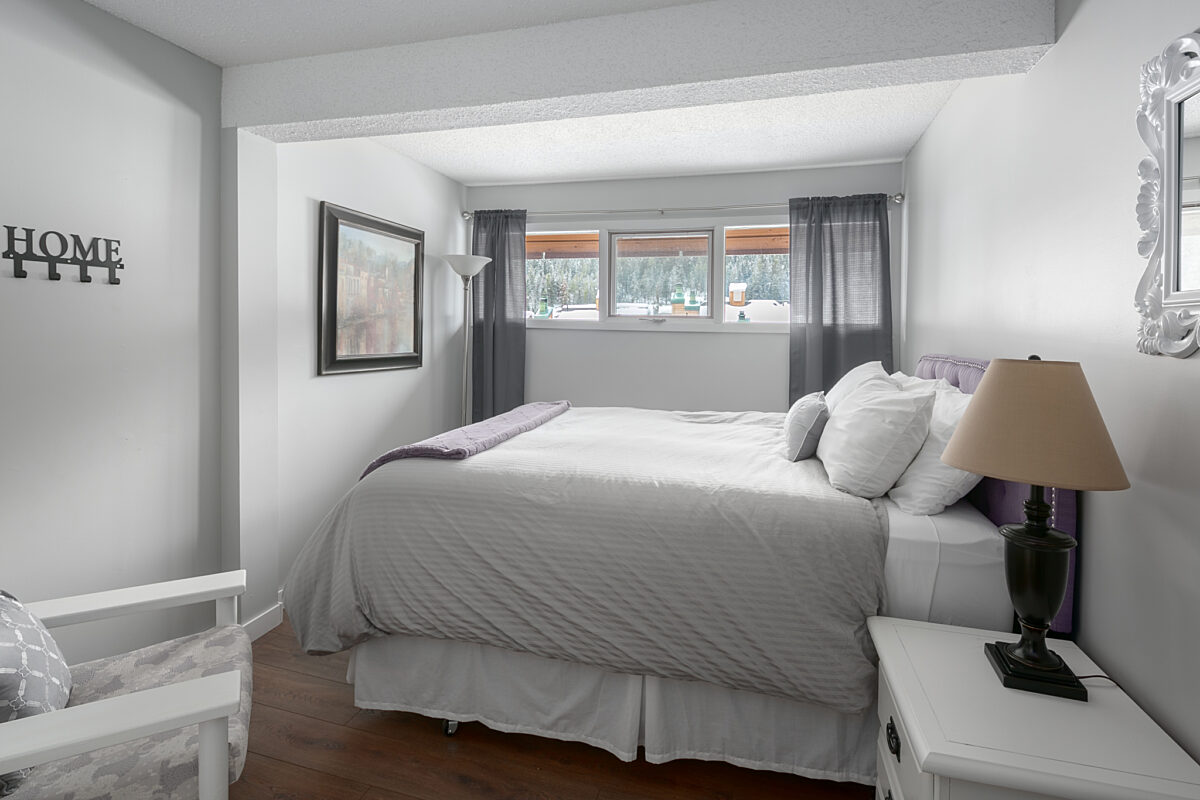 Queen bedroom with grey bedding, white pillows, curtains, and a chair to the left. Nightstand with lamp to the right.