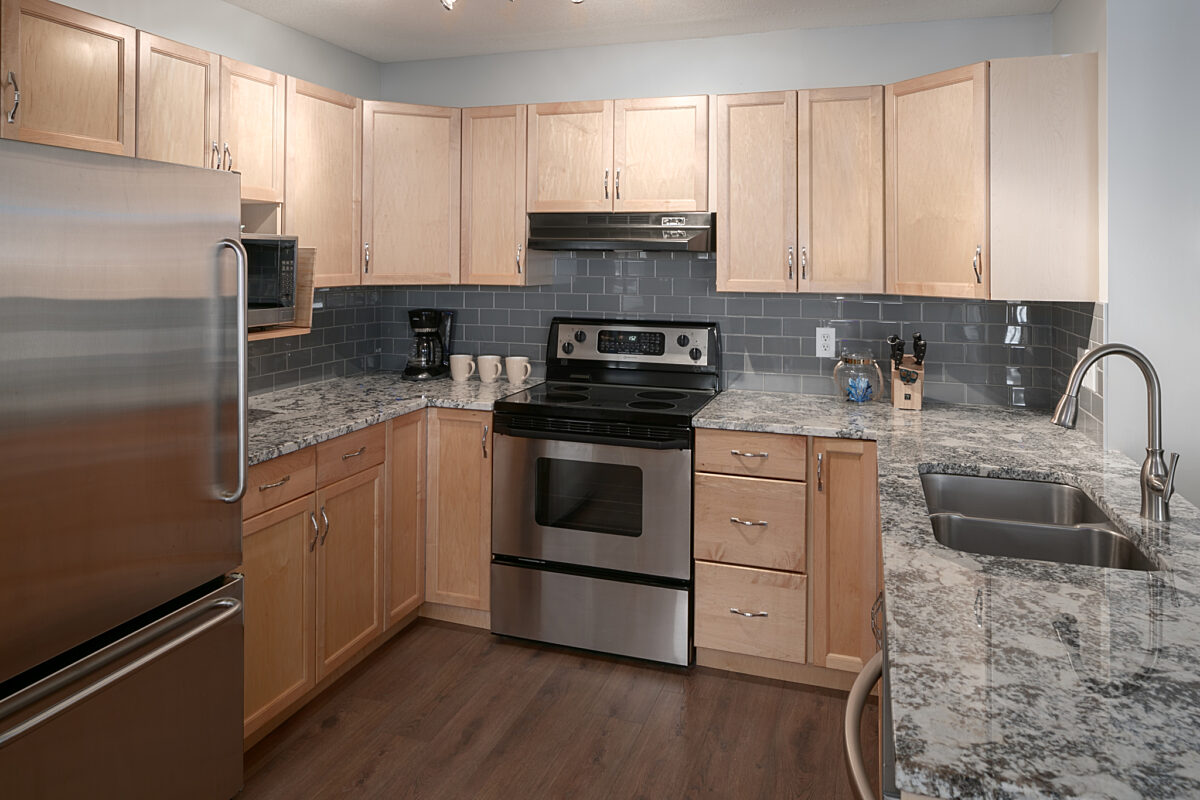 Kitchen area with light wood cabinets, stainless steel fridge, oven, and granite counterspace.