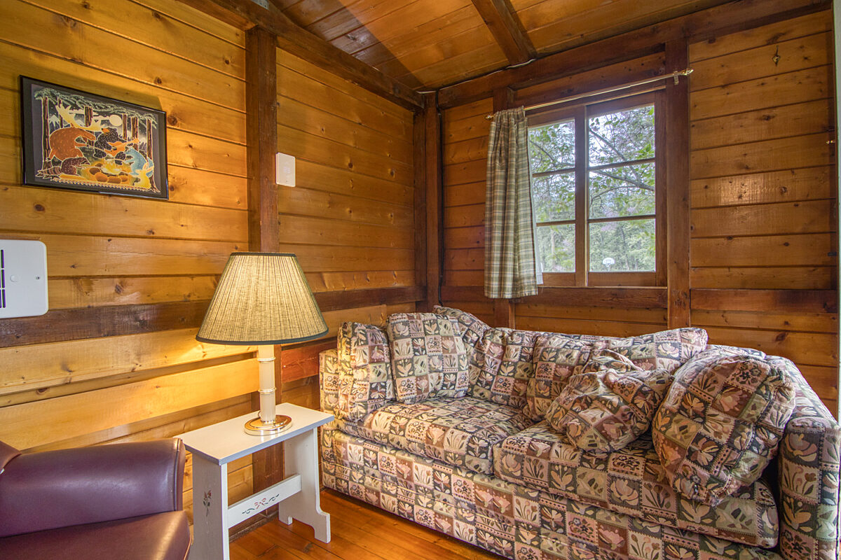 Living room of wood panelled cabin with floral couch, lamp, and window.