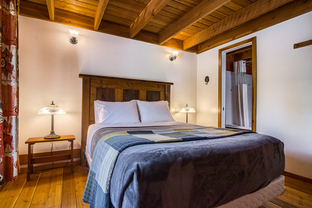 Queen bed with blue bedding, white pillows, and white walls and wood panelled ceiling.