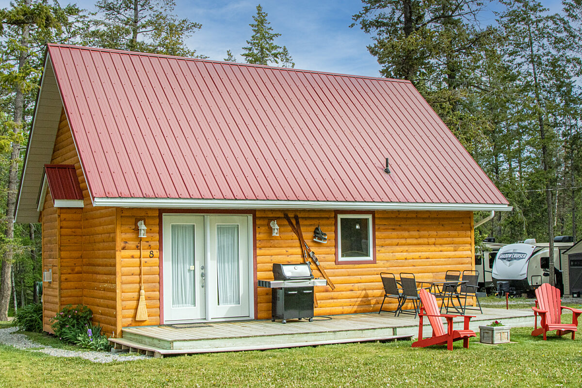 Wood cabin with red roof and white doors. BBQ on the patio and red chairs outside. Green grass outside.