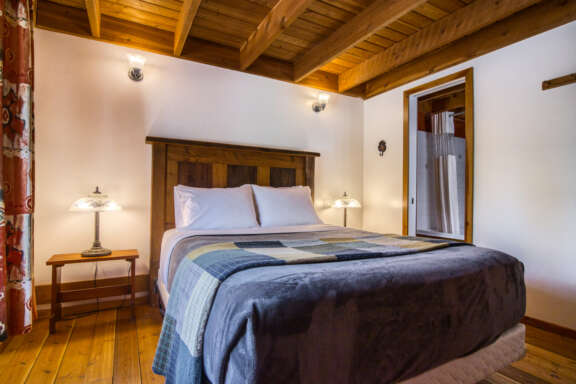 Queen bed with blue bedding, white pillows, and white walls and wood panelled ceiling.