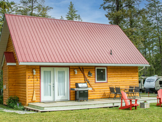 Wood cabin with red roof and white doors. BBQ on the patio and red chairs outside. Green grass outside.