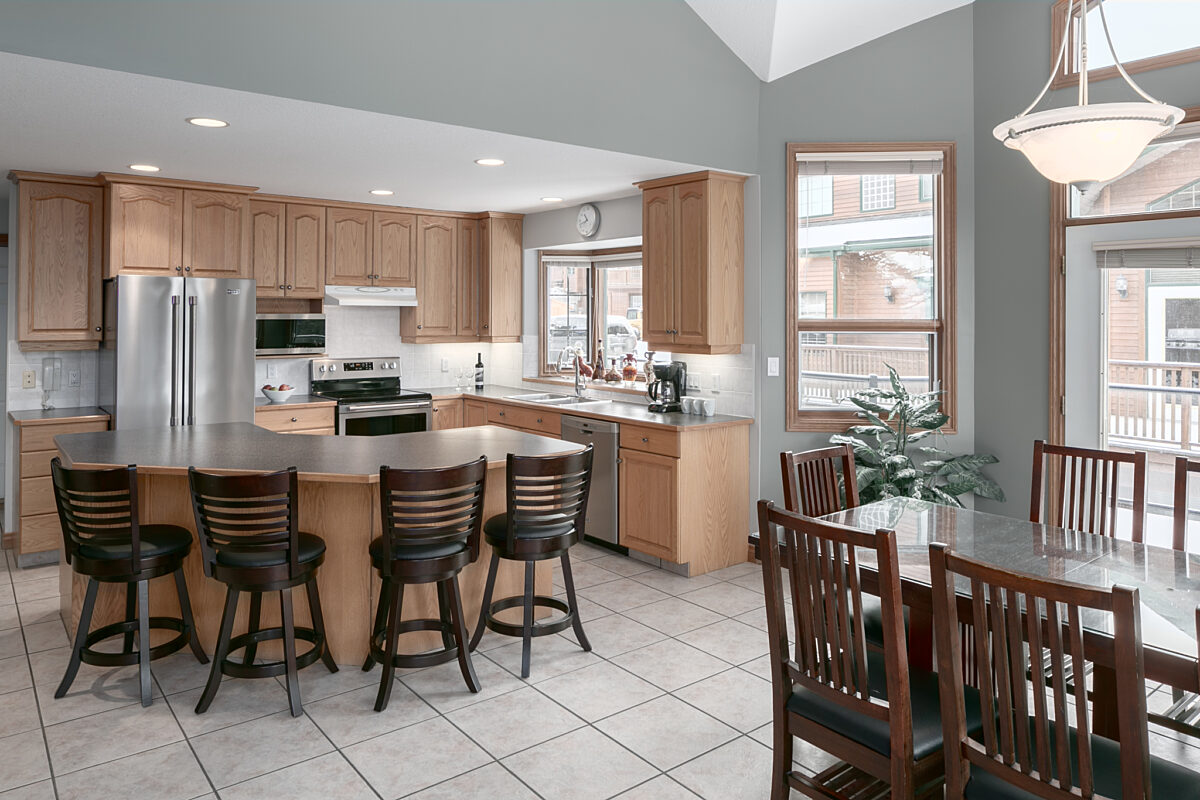 Kitchen table with four chairs to the right, kitchen island with four barstools to the left. Kitchen appliances in background.