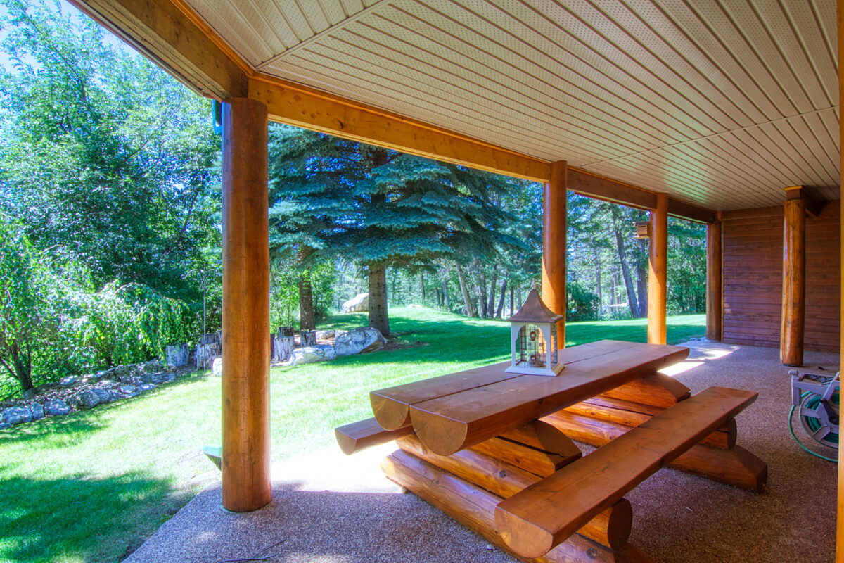 Log picnic table under an enclosed deck. Paved pad leads on to greenspace, grass, and treed area.