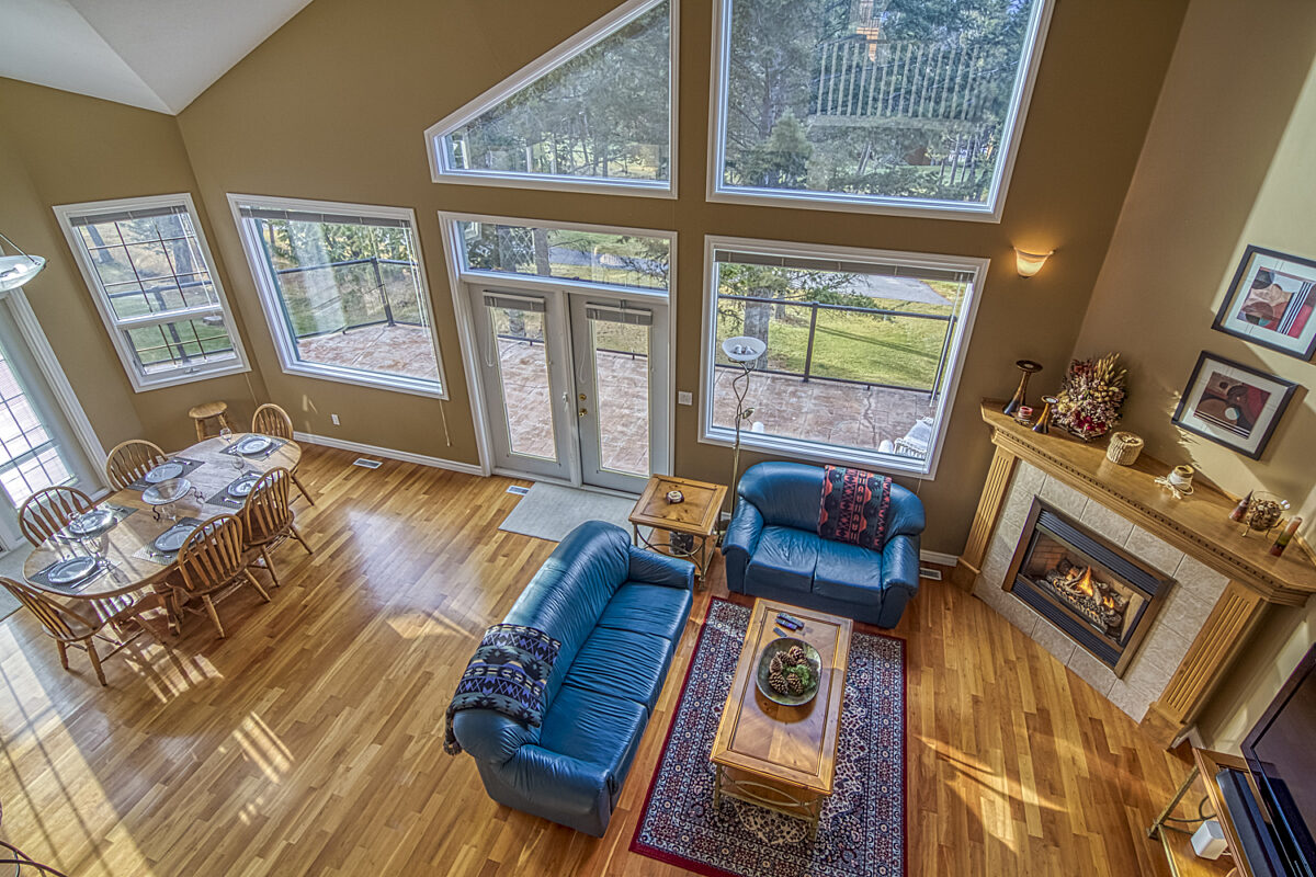 Dining room and living room as viewed from the loft above. Light wood dining table and chairs, blue couch and chair, rug, coffee table, and fireplace.