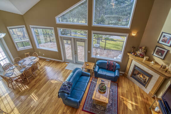 Dining room and living room as viewed from the loft above. Light wood dining table and chairs, blue couch and chair, rug, coffee table, and fireplace.