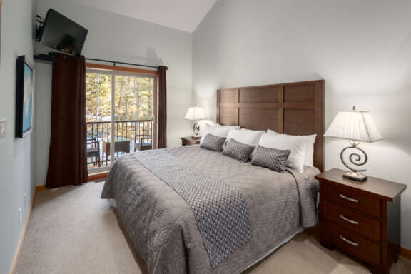 King bedroom with grey bedding, bedside tables and lamps on both sides of the bed. Windows to the left.