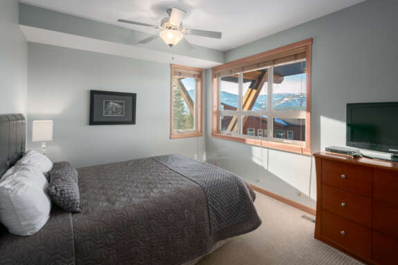 Queen bedroom with grey bedding, pillows, and window to the right. Dresser with television to the right.