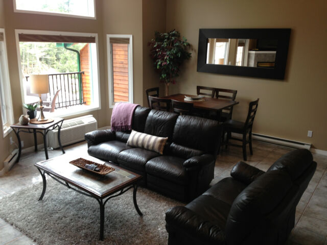 Spacious living room with black leather couches, and dining room table and chairs in background.