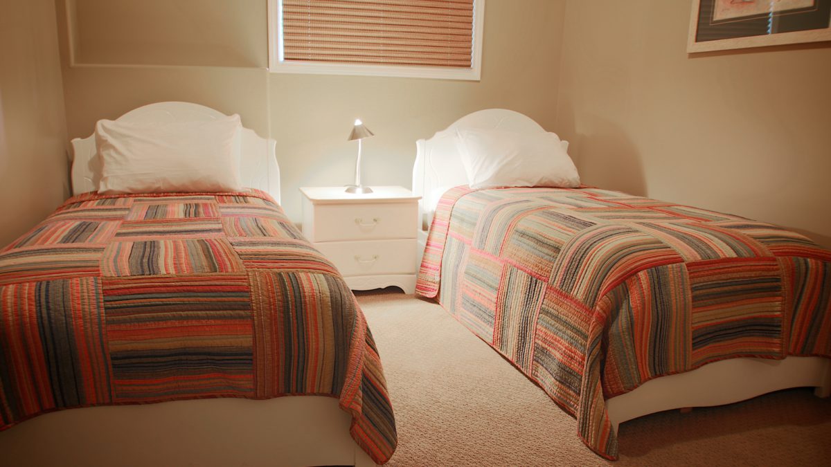 Bedroom with two twin beds, striped multicolour bedding, and bedside table with lamp in the centre.