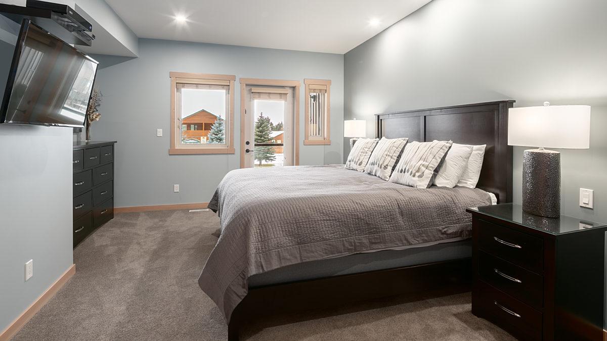 Queen bedroom with grey bedding, overhead lighting, two bedside tables, and windows to the left.