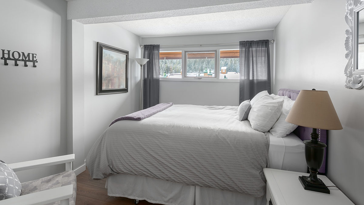 Queen bedroom with grey bedding, white pillows, curtains, and a chair to the left. Nightstand with lamp to the right.