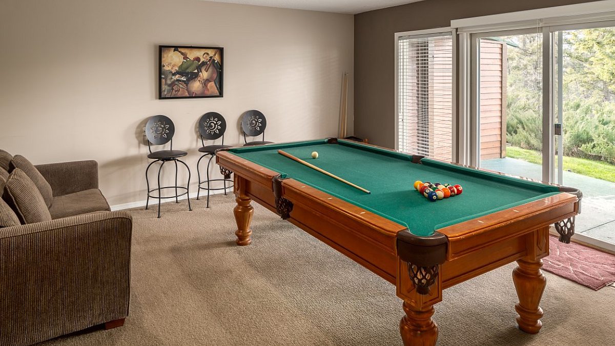 Pool table with balls and sticks. Couch and three barstools. To the left, large glass doors.