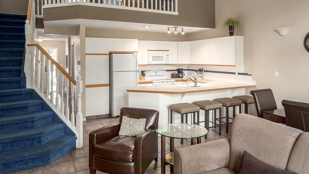 Kitchen and living space. Couch, chairs, and barstools at a kitchen island. To the right, blue carpeted stairs lead to the upper level of the suite.