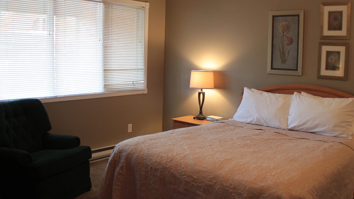 Master bedroom with queen bed. Beige bedding, pillows, bedside table, and lamp. Black fabric chair.