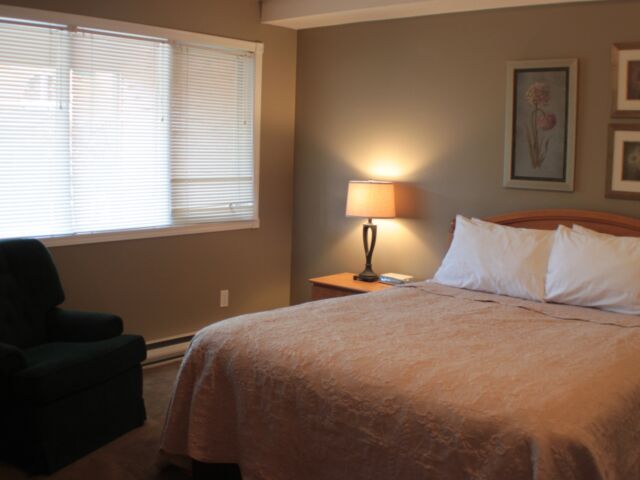 Master bedroom with queen bed. Beige bedding, pillows, bedside table, and lamp. Black fabric chair.