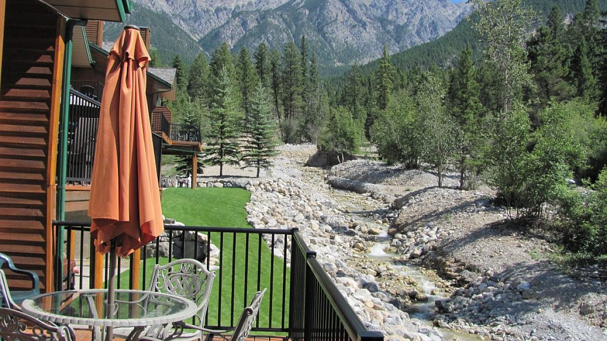 Patio with umbrella, table, and chairs. Overlooking a creek bed, trees, greenery, and mountains in the distance.