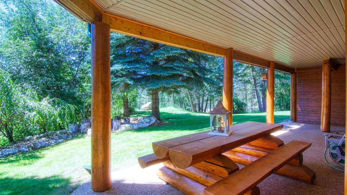 Log picnic table under an enclosed deck. Paved pad leads on to greenspace, grass, and treed area.