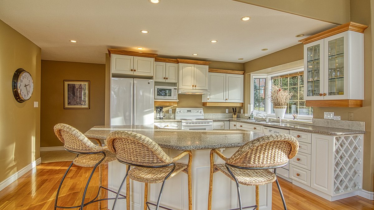 Spacious kitchen and kitchen island with light wood and white detailing. Three barstools.